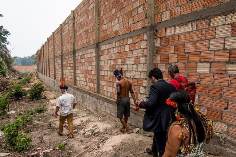 A imagem mostra cinco pessoas caminhando ao lado de um muro de tijolos em área de vegetação recém-derrubada. Três dessas pessoas usam trajes tradicionais e cocares indígenas.