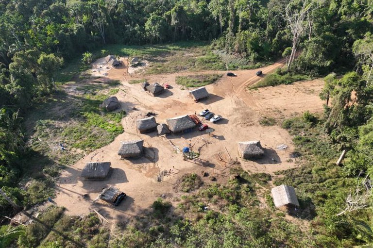 Foto em vista aérea de uma pequena aldeia Parakanã em uma clareira na floresta. A aldeia possui cabanas com telhados de palha. Há veículos estacionados em um terreno de terra batida.