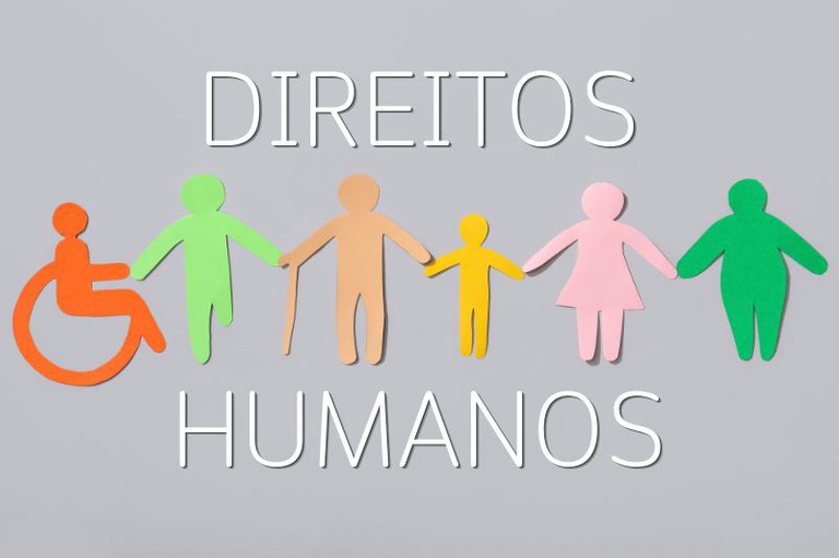 Recortes de papel coloridos em forma de pessoas diversas dão as mãos entre as palavras Direitos Humanos sobre um fundo cinza.