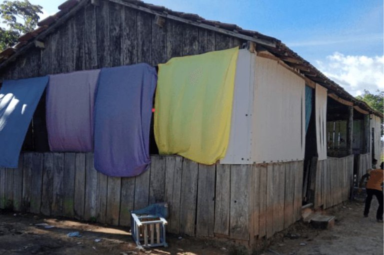 Imagem externa de uma escola construída em madeira. As janelas são tampadas por lençóis coloridos (azul, lilás e amarelo). O chão ao redor é de terra e, ao fundo, há vegetação sob um céu azul com algumas nuvens. Uma pessoa é parcialmente visível à direita.