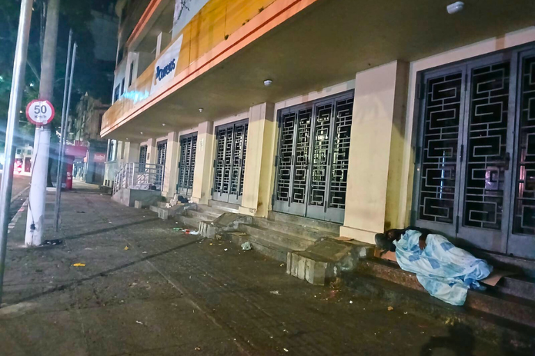 #ParaTodosVerem: Foto noturna da fachada de uma agência dos Correios com portas pantográficas fechadas. À direita, uma pessoa dorme sobre degraus, coberta por um cobertor azul e branco.