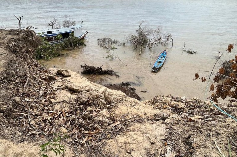 Margem de rio com terra desmoronada, árvores secas na água barrenta e dois barcos pequenos atracados sob o barranco íngreme de terra e galhos.