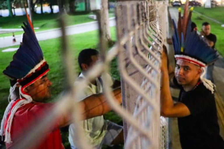 Foto em formato retangular de dois homens que usam cocares indígenas e estão ao lado de uma grade de metal. Um deles veste uma camisa vermelha, enquanto o outro usa uma camisa preta e olha para a câmera. O fundo inclui uma área verde iluminada e outras pessoas ao longe.