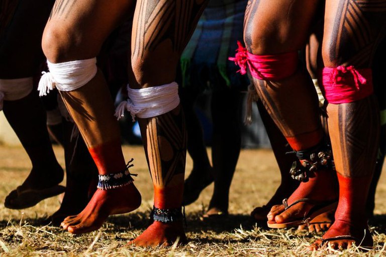 Foto de pernas de um grupo de pessoas. As pernas estão pintadas de vermelho e com urucum. Algumas pessoas usam faixas brancas ou cor-de-rosa amarradas nas panturrilhas e tornozeleiras com contas ou sinos. Os pés estão descalços ou calçados com sandálias, e eles estão em uma superfície com grama seca.