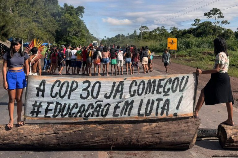 Foto em formato retangular de um grupo de pessoas bloqueando uma estrada com um tronco de árvore. Elas seguram uma faixa com a mensagem: "A COP 30 já começou #Educação em luta!".