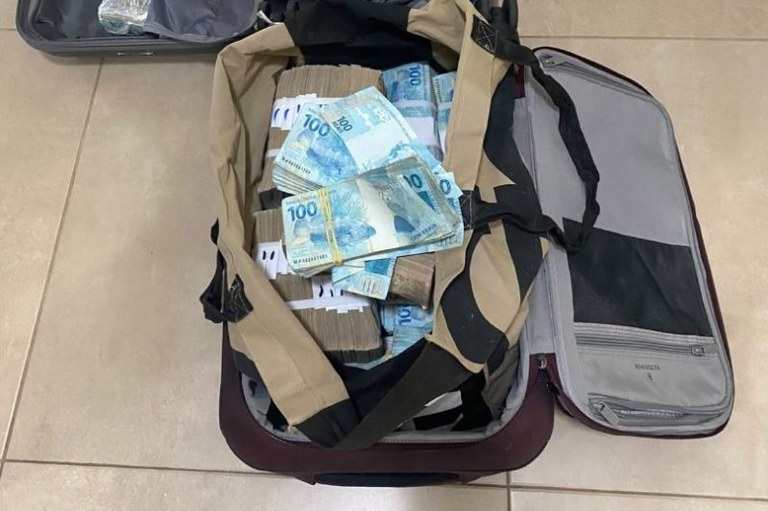 Foto em ângulo superior de uma mala aberta cheia de maços de notas de R$ 100 amarradas com elásticos e faixas de papel.
