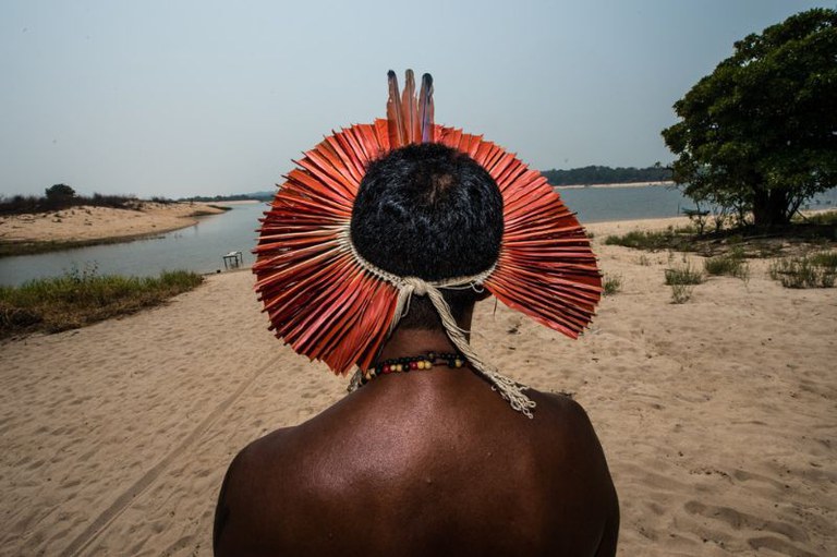 Foto de homem indígena visto de costas, usando um grande cocar circular avermelhado em uma praia de rio.