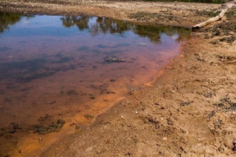 margem de rio com sedimento de cor laranja avermelhada, nitidamente contendo material ferroso ou laterítico.