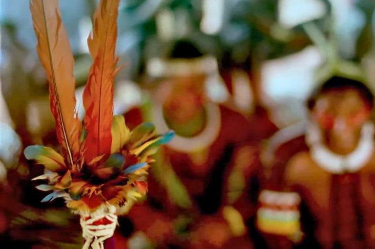 Foto em close-up de um adorno de penas coloridas, com indígenas em trajes tradicionais desfocados ao fundo.