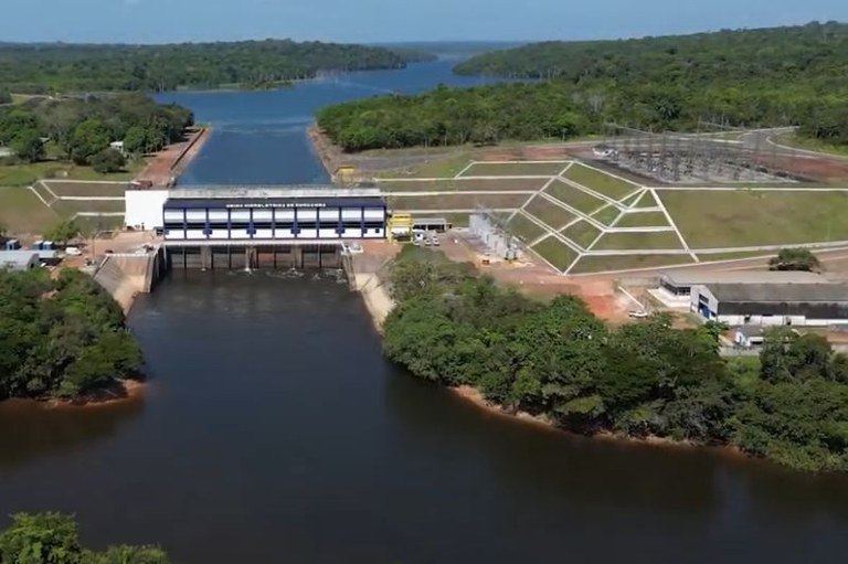 A imagem mostra a barragem principal com a casa de força, o reservatório de água a montante (acima), o rio a jusante (abaixo), e estruturas auxiliares como a subestação de energia. A usina está localizada em uma área de mata densa.