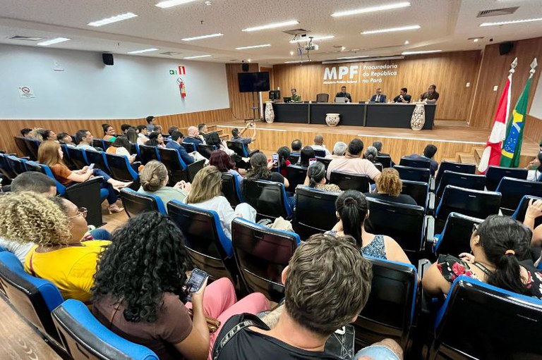 Auditório cheio com diversas pessoas sentadas em poltronas azuis, voltadas para uma mesa no palco com palestrantes e ao fundo o logotipo do MPF.