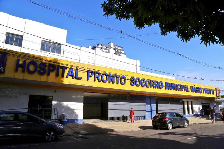 Foto da fachada do Hospital Pronto Socorro Municipal Mário Pinotti, em Belém. O prédio é branco com uma grande placa amarela na entrada, onde o nome do hospital está escrito em letras azuis. A imagem foi tirada da rua em um dia ensolarado.