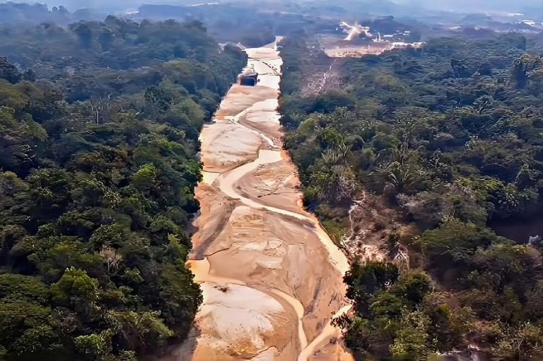Foto mostra uma área de floresta densa com um rio de baixo nível d'água, revelando grandes bancos de areia. A floresta é verde e densa, enquanto o leito do rio é predominantemente arenoso.