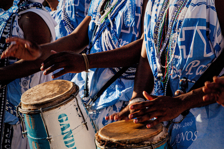 A imagem mostra um grupo de pessoas tocando tambores, vestindo roupas azul e branca com padrões geométricos e inscrições, além de colares de contas coloridas. Os braços e mãos dos músicos estão em movimento, batendo nos tambores. Os tambores são de madeira com pele esticada na parte superior, um deles tem a palavra "GAND" visível na lateral. A luz do sol ilumina a cena, criando sombras e destacando os detalhes das roupas e dos instrumentos.