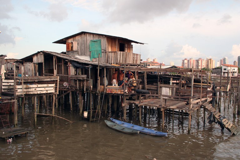 Palafitas de madeira sobre rio barrento com duas canoas ancoradas. Pessoas estão no deck de uma casa. Ao fundo, prédios urbanos sob céu nublado.