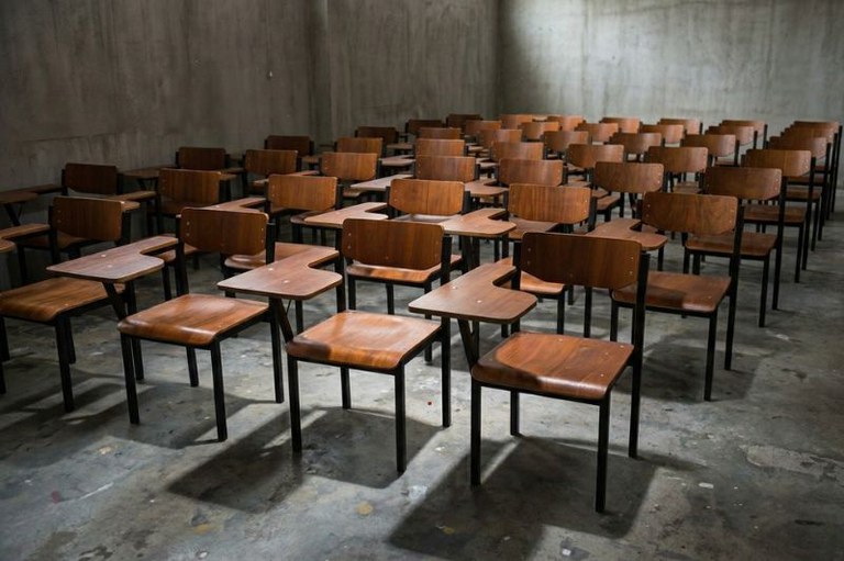 Sala com várias fileiras de cadeiras escolares de madeira e metal sobre piso de concreto e paredes cinzas.