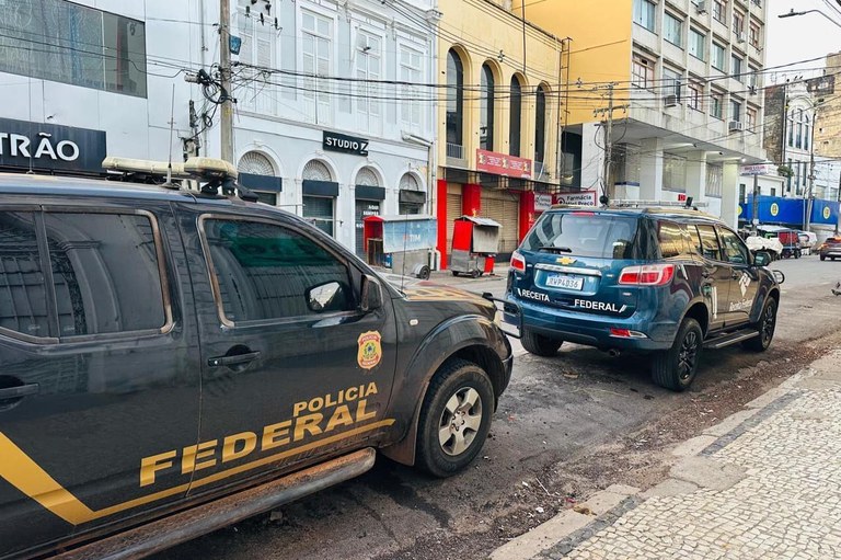 A imagem mostra duas viaturas da Polícia Federal e da Receita Federal paradas em uma rua de paralelepípedos. A viatura da Polícia Federal, um veículo preto com o nome "POLÍCIA FEDERAL" em amarelo na lateral, está em primeiro plano e parcialmente visível. A viatura da Receita Federal, um SUV azul com a inscrição "RECEITA FEDERAL" na parte traseira, está estacionada logo atrás. Ao fundo, há edifícios de dois andares, com lojas e fachadas de estilo antigo, e a rua parece ser uma via urbana.