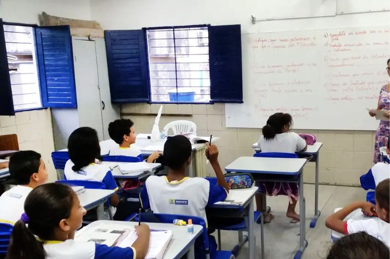 Foto de uma sala de aula de uma escola pública com crianças sentadas em suas carteiras, vestindo uniformes brancos e azuis, enquanto uma professora explica o conteúdo escrito no quadro branco ao fundo.