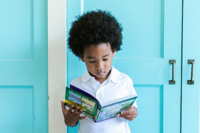 Foto de criança que lê livro colorido enquanto está em frente a uma porta azul clara. A criança veste uma camisa branca.