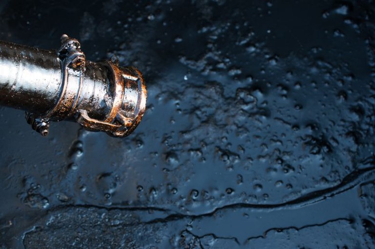 Foto retangular em close-up de um bocal de mangueira metálico coberto por petróleo, que forma uma poça espessa e irregular sobre uma superfície escura abaixo.