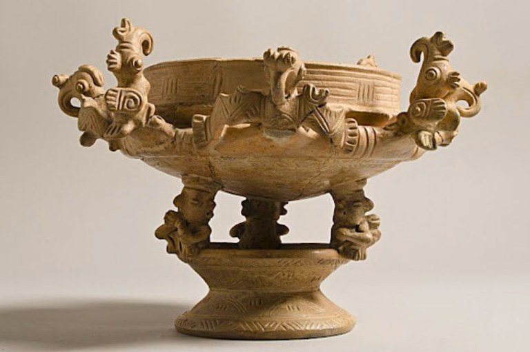 Vaso de cerâmica bege sobre fundo neutro. Três figuras de base sustentam a tigela, decorada com relevos e esculturas de pássaros na borda.