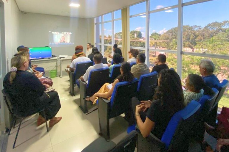 Foto interna de uma sala com um grupo de pessoas sentadas em cadeiras, assistindo a uma apresentação. Uma tela exibe a imagem de uma pessoa em videoconferência. A sala possui grandes janelas à direita, com vista para árvores e céu azul
