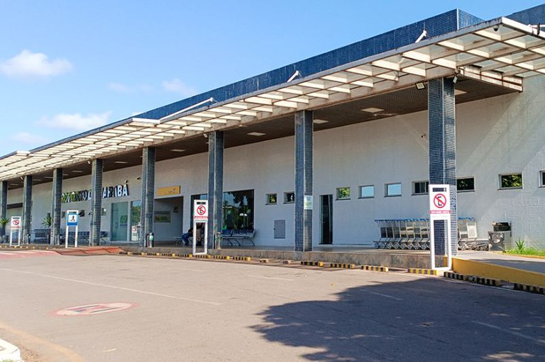 Foto da fachada do aeroporto de Marabá, com uma longa cobertura sustentada por pilares revestidos de azulejos escuros. A parede principal do edifício é branca, com janelas retangulares. À esquerda, há sinalização indicando o nome do aeroporto. Em primeiro plano, uma via de acesso com meio-fio pintado de amarelo e preto, e alguns carrinhos de bagagem. O céu está azul e ensolarado.