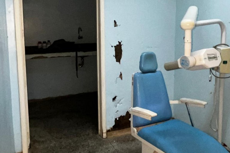 Foto de consultório odontológico em péssimas condições, com cadeira azul desgastada, paredes danificadas, mofo e infiltrações. Ao fundo, sala escura com pia quebrada e ambiente deteriorado.