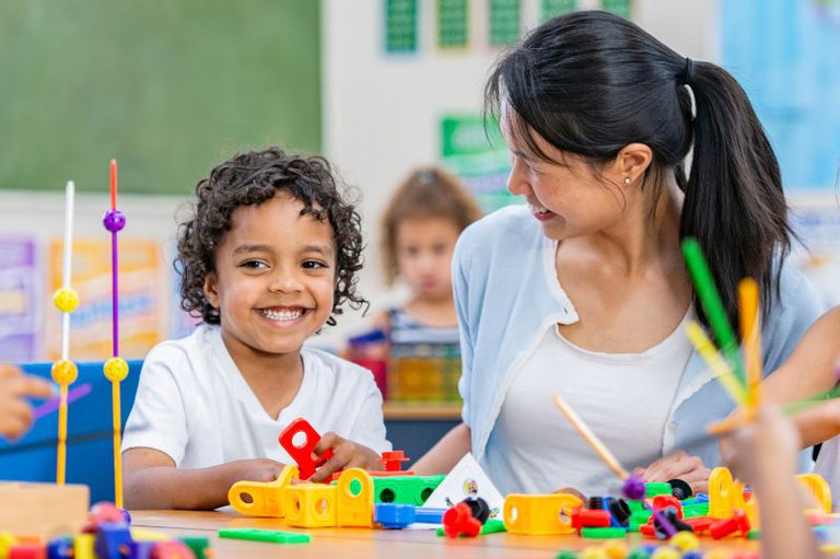 Uma pessoa adulta de cabelo preto amarrado está sorrindo para um menino negro com cabelos encaracolados que está sorrindo de volta. Ambos estão sentados a uma mesa de madeira e brincando com brinquedos de construção coloridos. Há outras crianças em segundo plano, ligeiramente desfocadas.