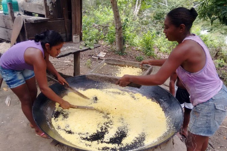 Duas mulheres em ambiente externo processam farinha amarela em um grande tacho circular de metal preto usando peneira e espátulas de madeira.