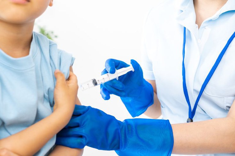 Foto de mãos com luvas azuis que aplicam uma injeção no braço de uma criança, que veste uma camiseta azul.