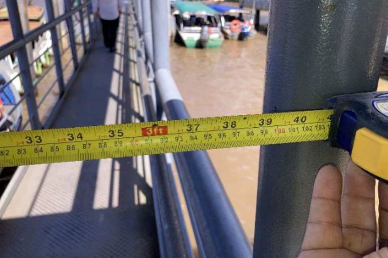 Foto diurna de mãos de pessoa que segura fita métrica amarela e preta, para medir entre estrutura metálica no terminal hidroviário Ruy Barata, em Belém. Ao fundo, passarela gradeada com pessoa caminhando, água marrom turva e barcos coloridos desfocados.