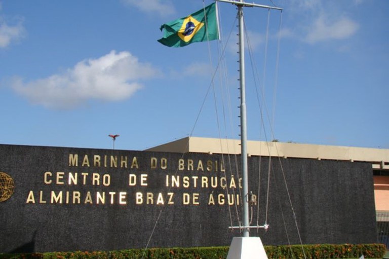 Foto da fachada do Centro de Instrução Almirante Braz de Aguiar, com muro preto com o nome da instituição escrito em letras de metal dourado. Há uma bandeira do Brasil hasteada em um mastro à frente.
