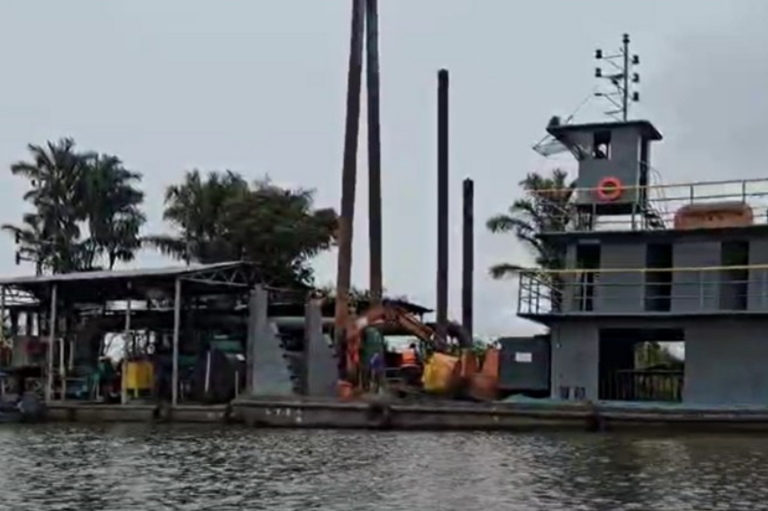 Foto de uma balsa de dragagem de cor predominantemente cinza que opera no rio Tapajós. Na embarcação, há altos postes verticais (estacas de fixação), uma escavadeira com braço laranja em atividade e uma estrutura coberta com maquinário. Trabalhadores com coletes de segurança estão presentes. Ao fundo, há vegetação densa, incluindo palmeiras, sob um céu nublado.