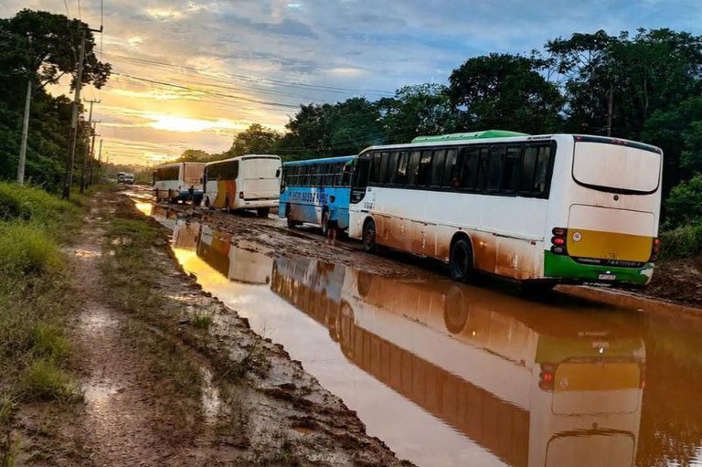 Quatro ônibus alinhados em uma estrada de terra lamacenta e alagada. O reflexo dos veículos aparece na água ao lado da vegetação.