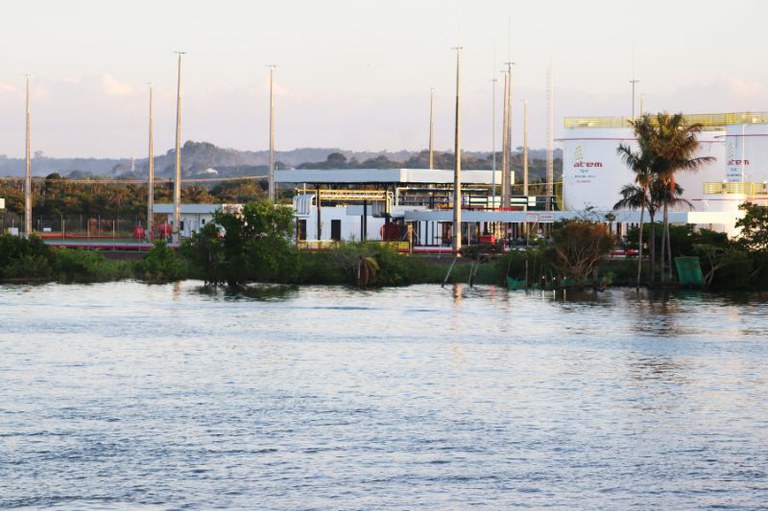 Vista de um corpo d'água, com vegetação na margem, e ao fundo, instalações industriais brancas com postes altos e um coqueiro.