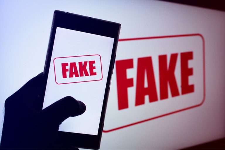 Foto de celular em cuja tela está escrito Fake. A palavra está na cor vermelha sobre fundo branco. O celular é segurado por pessoa em frente a tela maior. Nessa outra tela também está escrito Fake, nas mesmas cores.