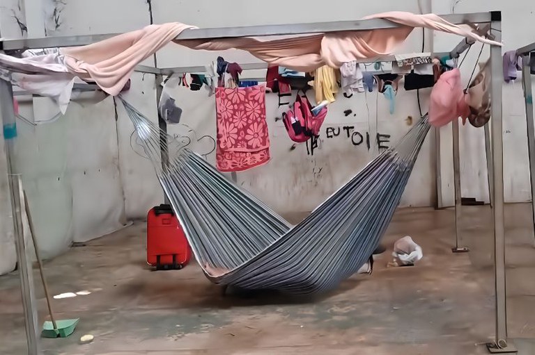Foto interna de galpão focado em estrutura metálica quadrada de onde pendem uma rede listrada cinza, roupas e toalha rosa.