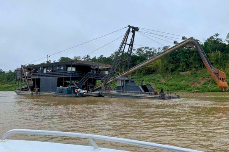 A imagem mostra uma grande embarcação metálica, própria para serviço de dragagem, em um rio barrento, equipada com um longo braço mecânico com uma concha na ponta. Há uma estrutura de dois andares, um pequeno barco ancorado ao lado e uma margem com vegetação densa ao fundo.