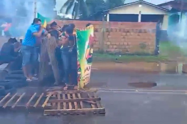 Foto de um grupo de pessoas protestando em uma via pública de área residencial. O grupo segura uma grande bandeira multicolorida, utilizando-a para se defender de balas de borracha. Pneus e paletes de madeira formam barricadas improvisadas, enquanto uma névoa de fumaça densa recobre o cenário ao fundo.