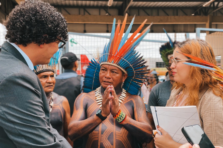 #ParaTodosVerem: a imagem mostra um procurador da República usando terno cinza, óculos e cabelo cacheado. Ao lado dele e centralizado na foto, há um homem indígena que usa um cocar azul e vermelho com pontas longas. O indígena está com as mãos juntas em frente ao corpo e possui pinturas corporais em tons escuros, cobrindo o peito e os braços, além de usar colares e pulseiras coloridas. Atrás dele, outro homem indígena com cocar semelhante observa a interação. Do lado direito da imagem, uma mulher com cabelos longos e claros, óculos, veste um blazer bege e segura documentos em uma mão enquanto olha para o indígena.