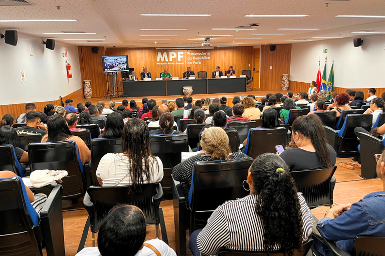 Ao fundo, mesa com cinco pessoas sob painel de madeira. à frente, público sentado de costas em poltronas pretas acompanha a reunião pública.