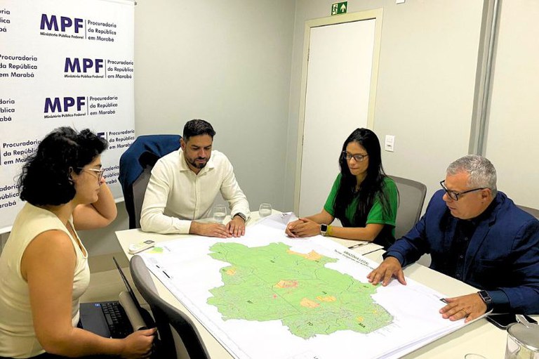 Foto de quatro pessoas reunidas em uma sala, em torno de uma mesa branca, analisando um grande mapa da Amazônia Legal. Uma mulher à esquerda consulta um laptop, enquanto um homem à direita gesticula sobre o mapa. Ao fundo, um banner do MPF - Procuradoria da República em Marabá.