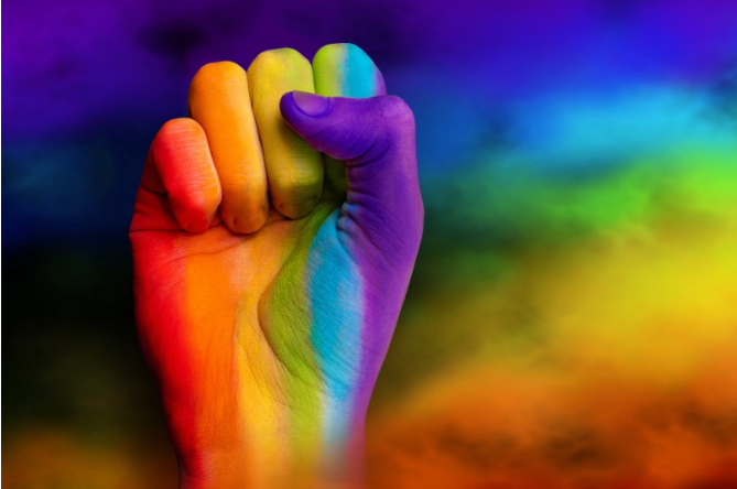 Fotografia ilustrativa mostra um punho fechado pintado com as cores do arco-íris, erguido à esquerda sobre um fundo nublado e desfocado com as mesmas cores, em referência às pessoas LGBTQIA+.