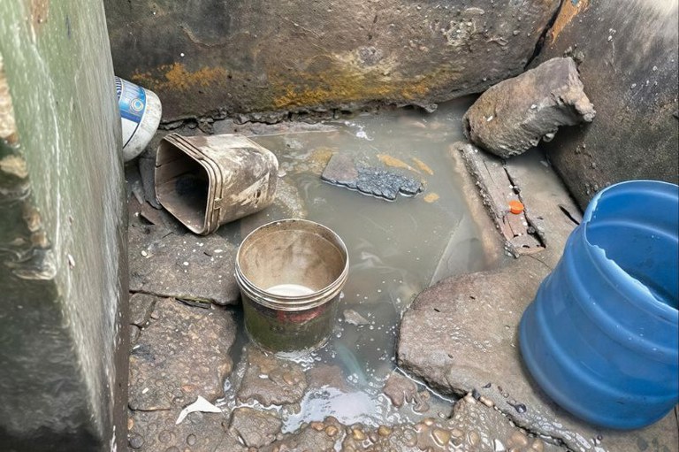 Recinto com paredes de pedra, poça d'água turva no chão, baldes de plástico sujos e restos de materiais de construção espalhados