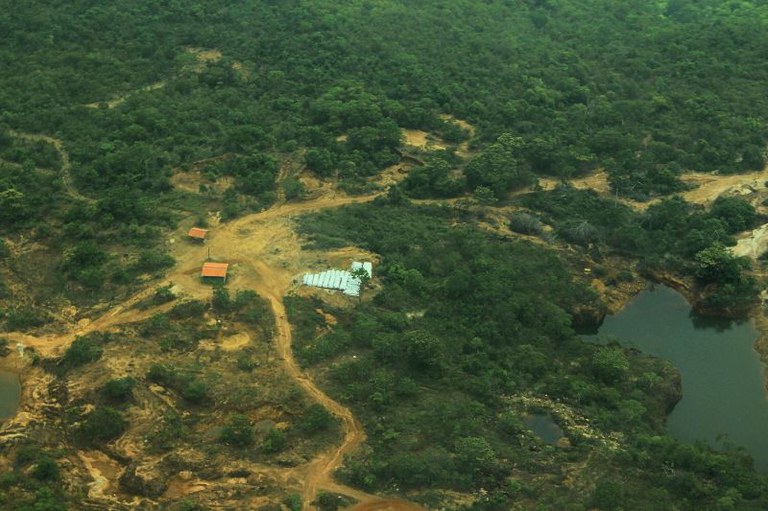 Vista aérea de uma área de mineração em uma floresta, mostrando clareiras de terra, pequenas construções e poços de água.