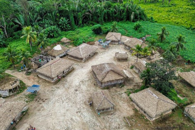 Vista aérea de uma aldeia indígena com várias habitações de palha dispersas sobre chão de terra clara, cercada por floresta verde.
