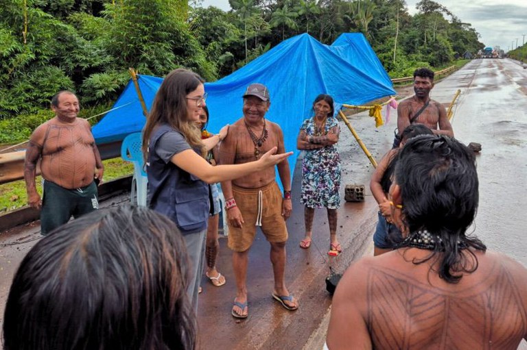 Foto da procuradora da República Thaís Medeiros da Costa de colete azul conversando com um grupo de indígenas com pinturas corporais tradicionais em uma estrada molhada. Há uma estrutura coberta com lona azul ao lado. Ao fundo vê-se floresta densa e alguns veículos bloqueados na estrada.