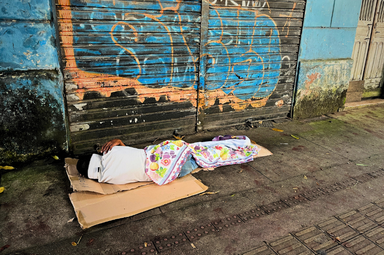 #ParaTodosVerem: Pessoa deitada sobre um papelão na calçada, coberta por um lençol estampado, em frente a uma porta de enrolar de metal com pichação azul e paredes desgastadas.