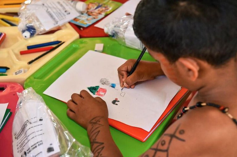 A imagem mostra uma criança desenhando em um papel com lápis de cor. O desenho inclui uma árvore, uma casa e outros objetos pequenos. A criança tem pinturas indígenas nos braços e usa um colar. A mesa está cheia de materiais de arte, incluindo lápis de cor, papel, bandejas de plástico e alguns materiais impressos.